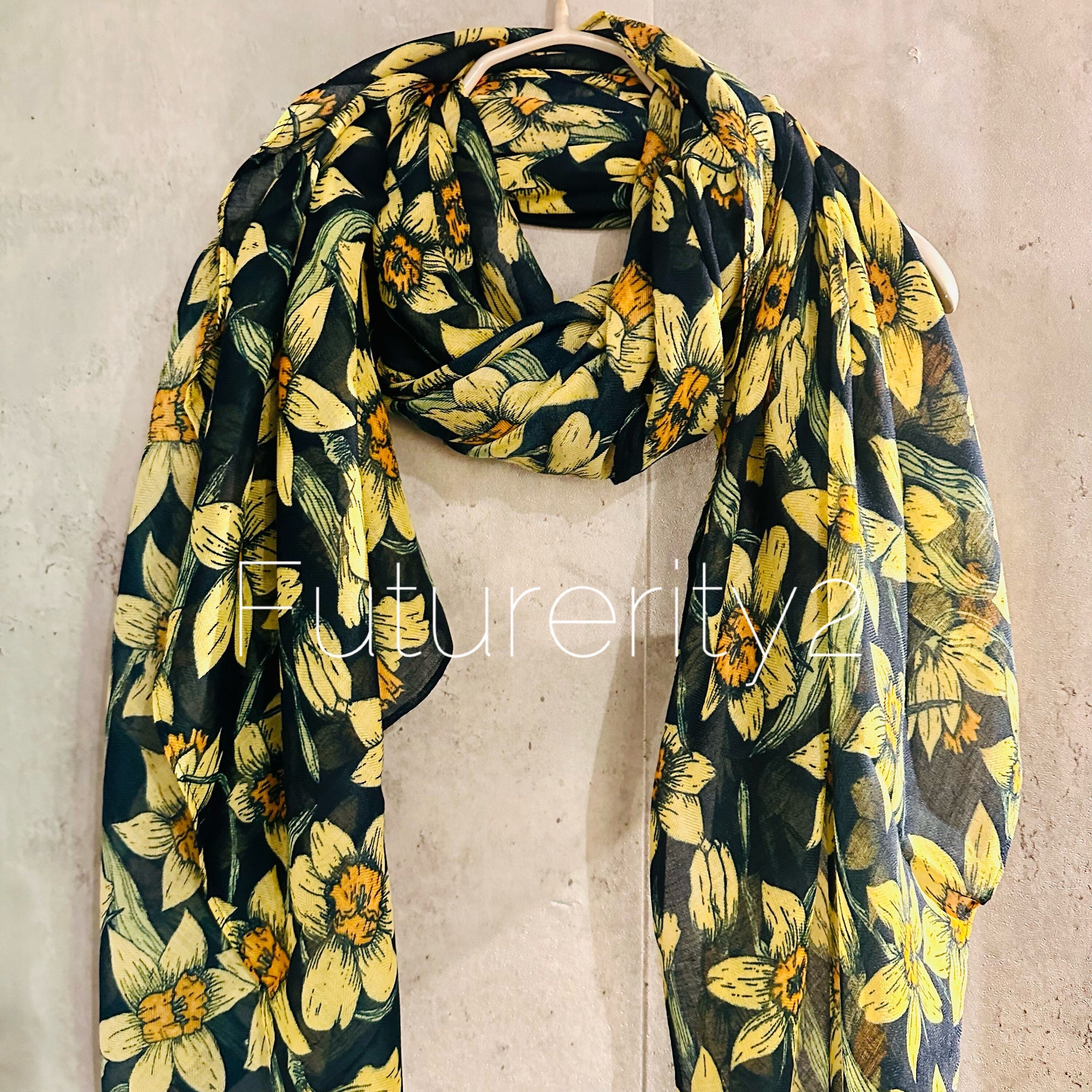 A black sheer scarf with a yellow floral print. The scarf is made of a lightweight fabric and has a flowing design. The scarf is displayed on a hanger against a textured gray wall.