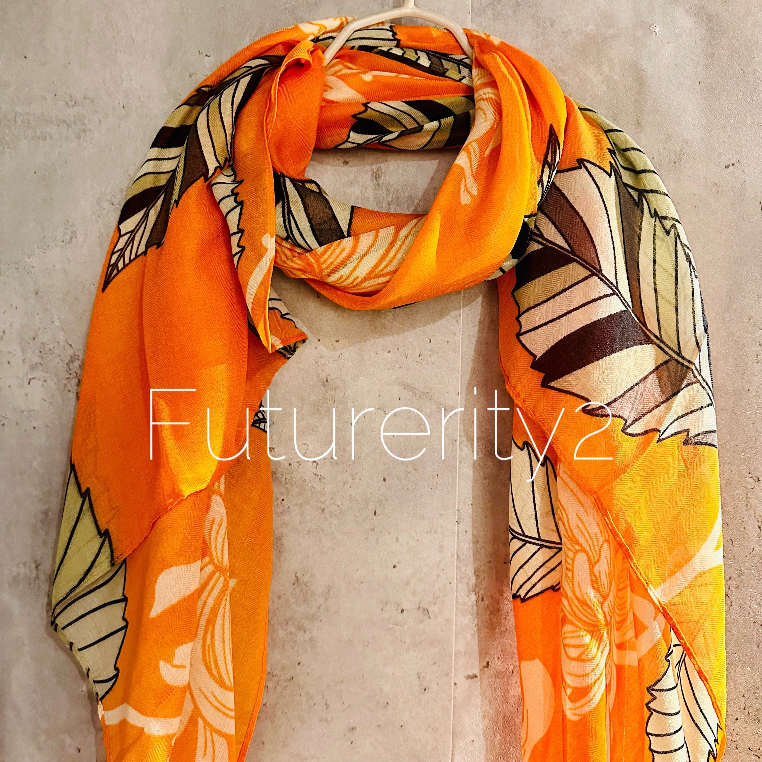 An orange scarf with a floral print. The flowers are orange and the leaves are green. The scarf is folded in half and draped over a hanger.