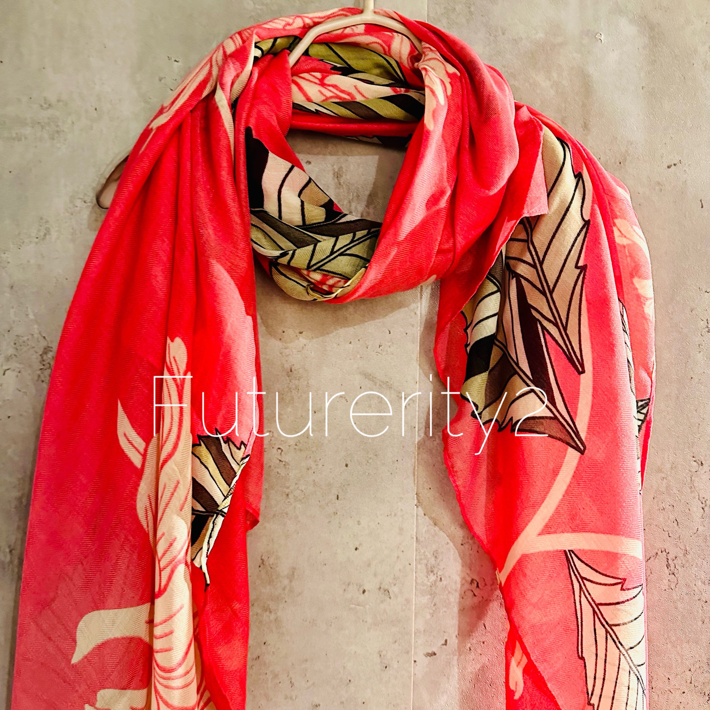 A pink scarf with a floral pattern. The pattern includes green leaves and white flowers. The scarf is made of a lightweight fabric and is draped over a hanger.