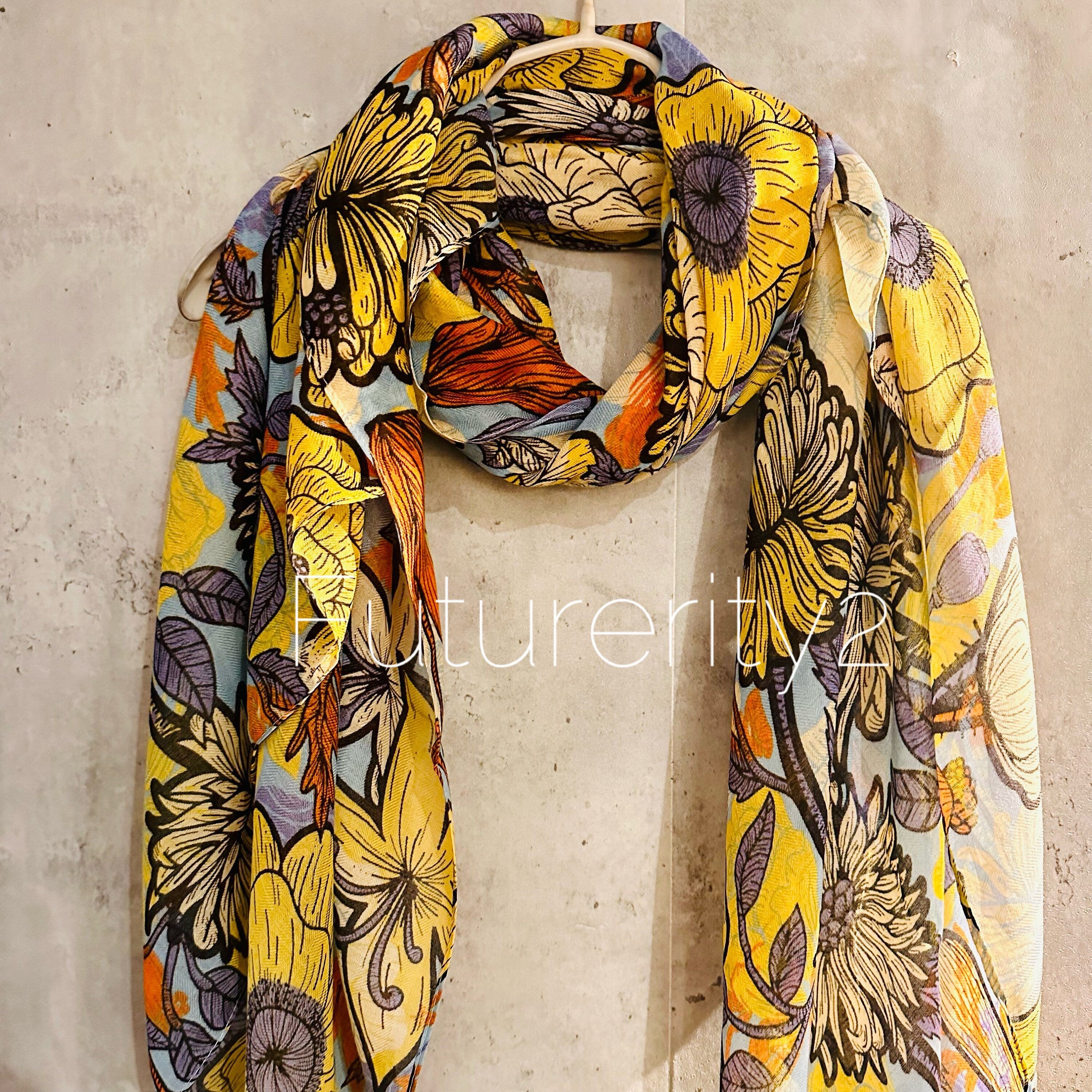 patterned scarf with a light blue background. The scarf is made of a soft, lightweight fabric and has a flowing design. The scarf is displayed on a hanger against a textured, gray background.